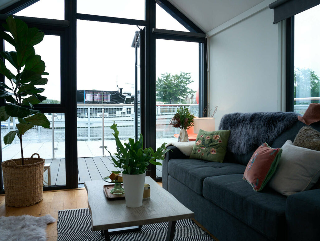Living area of houseboat showing sofa with cushions, coffee table, plants and view out over the marina through the floor to ceiling front windows